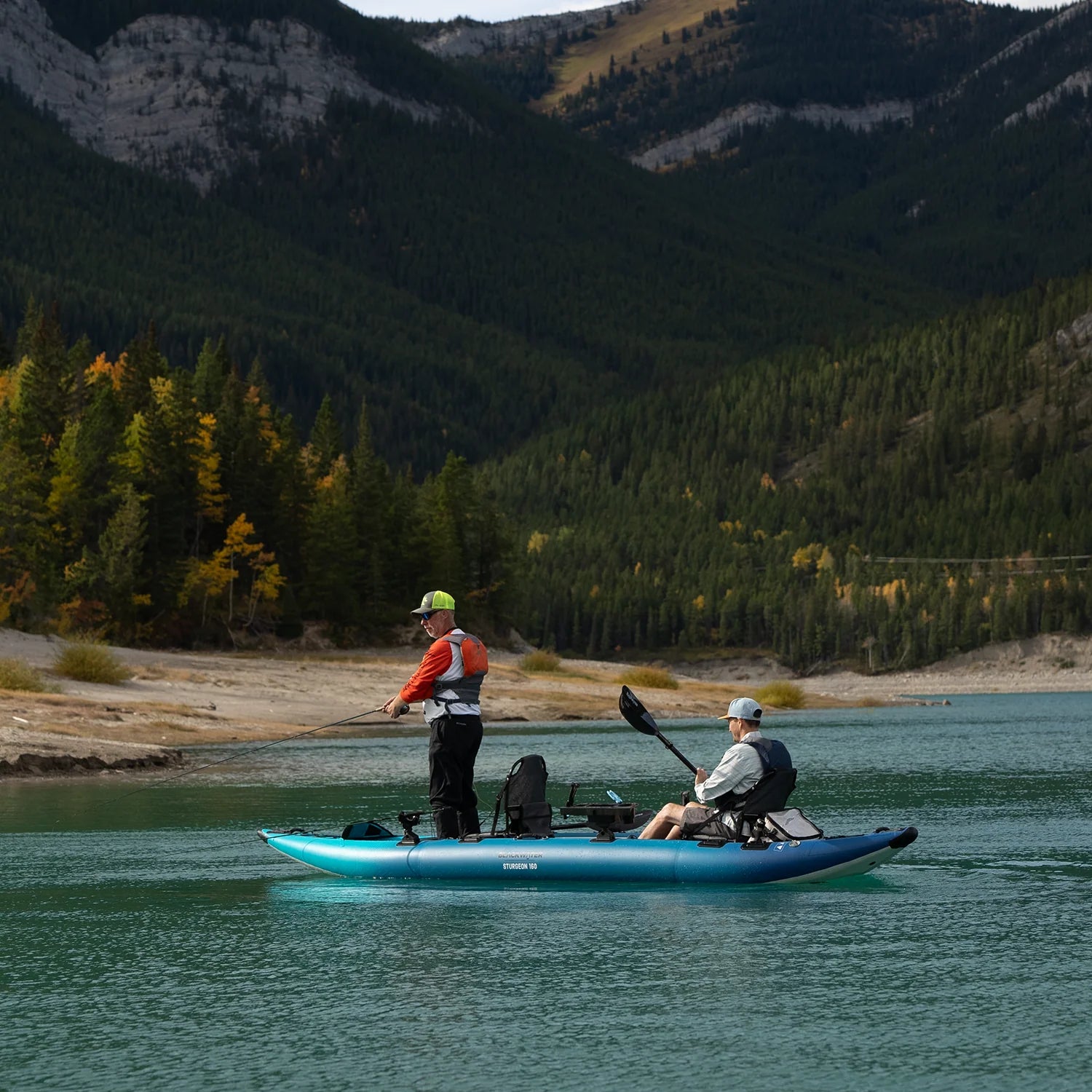 Two people on a blue inflatable kayak in a lake with mountains and trees in the background