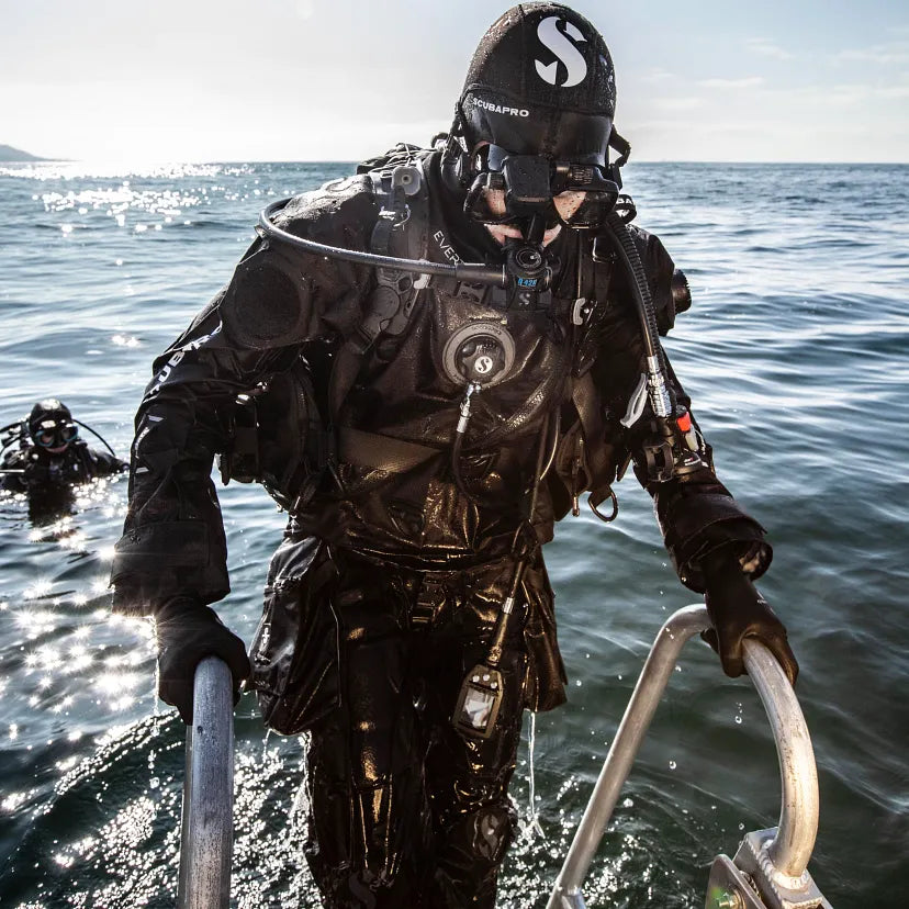 Person in a diving suit and helmet standing on a boat with water in the background