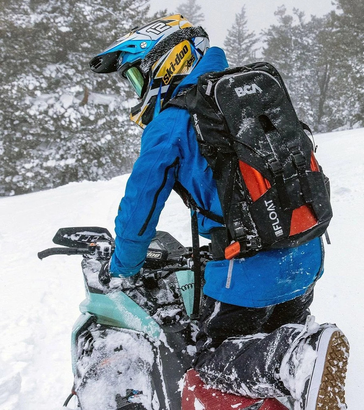 Person on a snowmobile wearing a blue jacket and helmet with a black backpack in a snowy landscape.