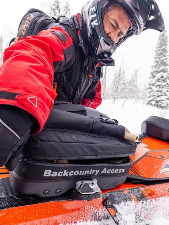 Person in winter gear sitting on a snowmobile with 'Backcountry Access' branding in a snowy landscape.